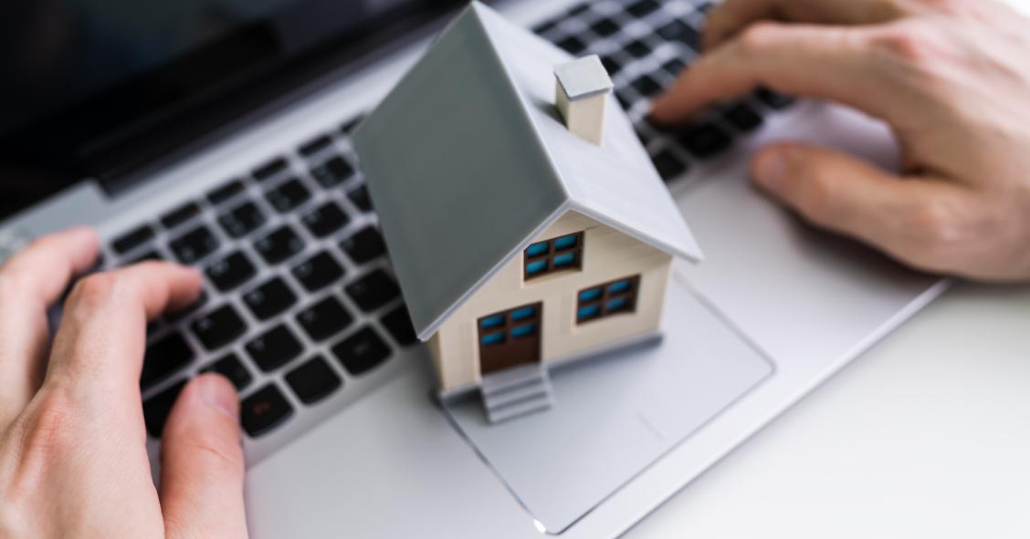 Mãos de uma pessoa a usar um computador portátil com uma miniatura de casa sobre o teclado, simbolizando crédito habitação online, simulação de empréstimo ou financiamento imobiliário digital.