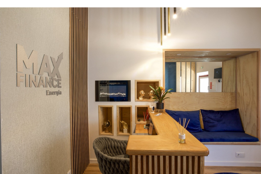 Sala de receção da MaxFinance Energia com balcão em madeira clara, parede com o logótipo prateado da empresa e zona de espera com almofadas azuis. O ambiente combina linhas modernas e iluminação suave, criando uma atmosfera acolhedora e profissional.