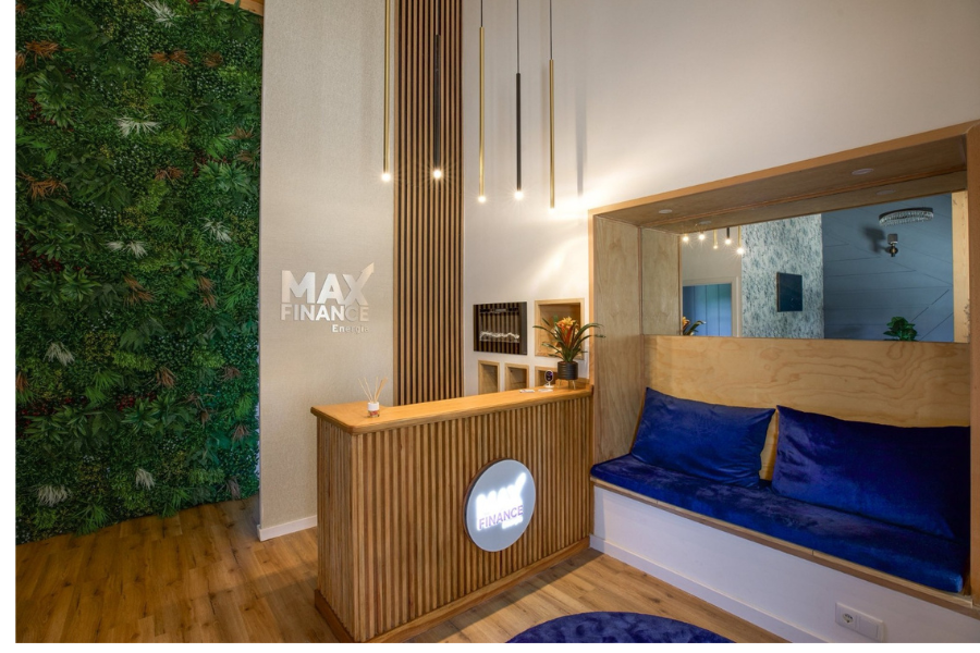 Vista ampla da receção da MaxFinance Energia, com balcão em madeira e logótipo iluminado, banco com almofadas azuis e parede lateral revestida de plantas verdes, simbolizando sustentabilidade e conforto. Iluminação pendente moderna e decoração minimalista completam o espaço.