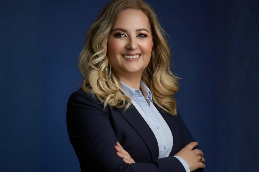Fotografia profissional de uma mulher de cabelo loiro e comprido, com blazer azul-escuro e camisa clara, de braços cruzados e sorriso confiante, sobre fundo azul.