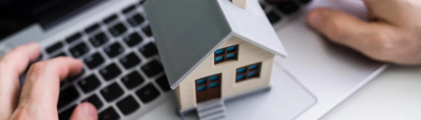 Mãos de uma pessoa a usar um computador portátil com uma miniatura de casa sobre o teclado, simbolizando crédito habitação online, simulação de empréstimo ou financiamento imobiliário digital.