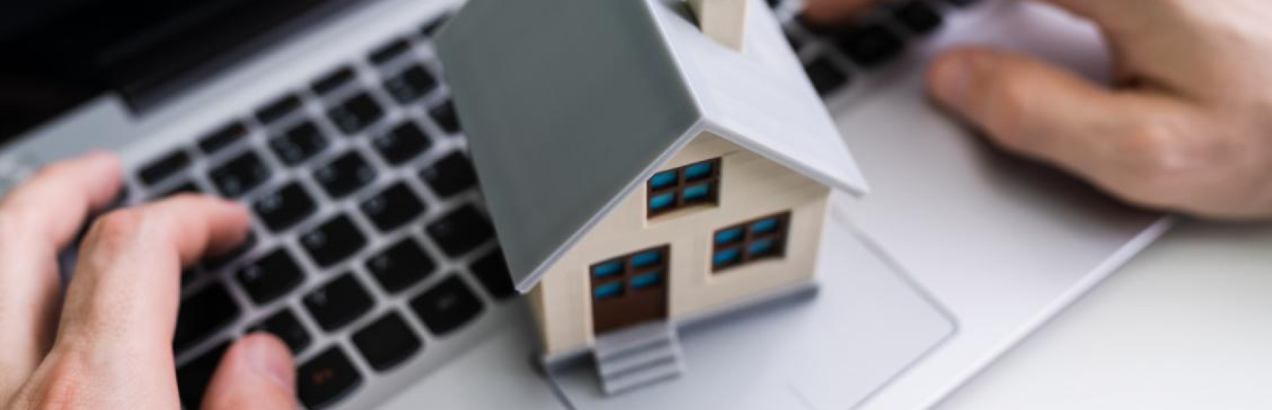 Mãos de uma pessoa a usar um computador portátil com uma miniatura de casa sobre o teclado, simbolizando crédito habitação online, simulação de empréstimo ou financiamento imobiliário digital.
